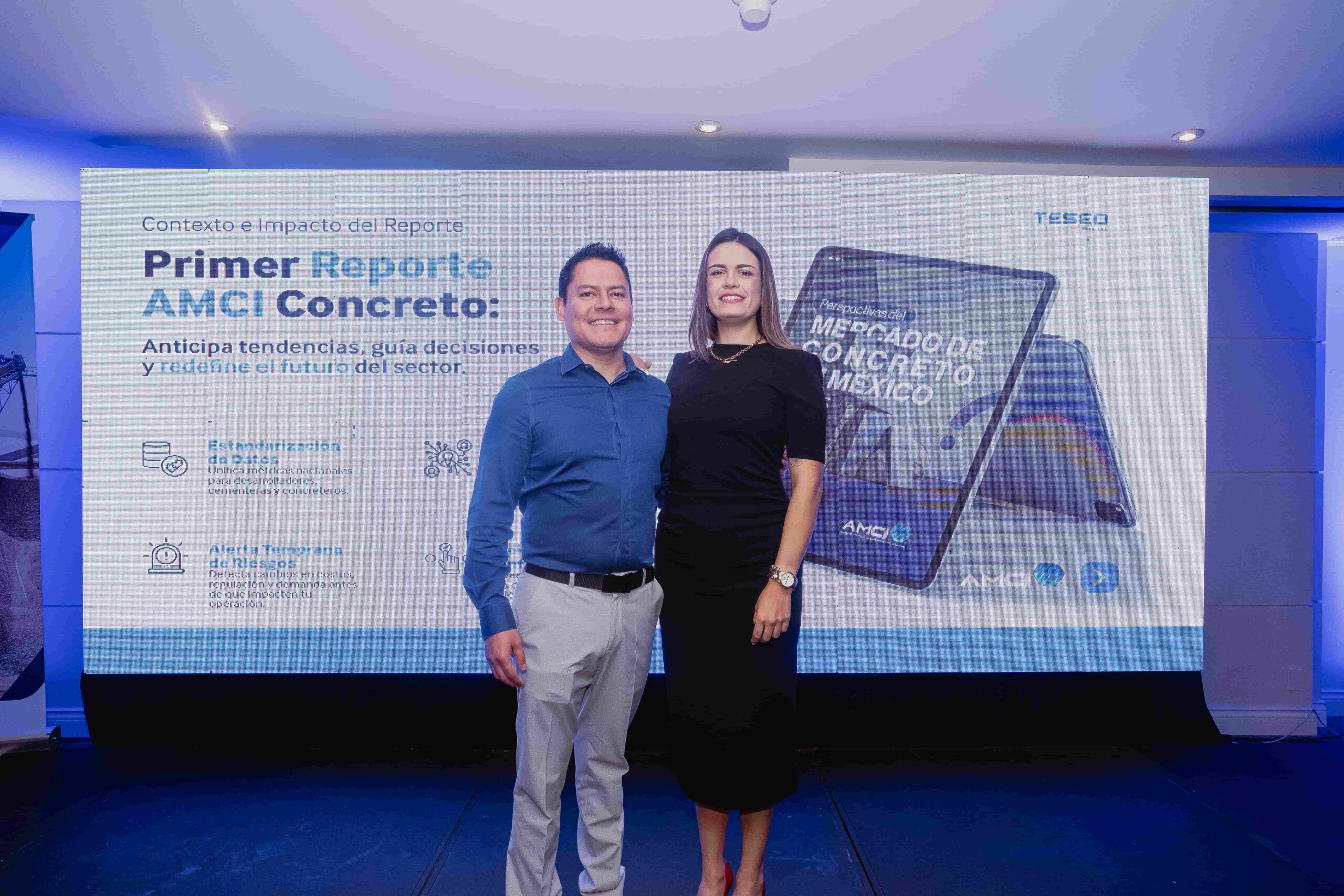 Dos personas posan frente a una pantalla con la diapositiva “Primer Reporte AMCI Concreto” de Teseo Data Lab durante la presentación.